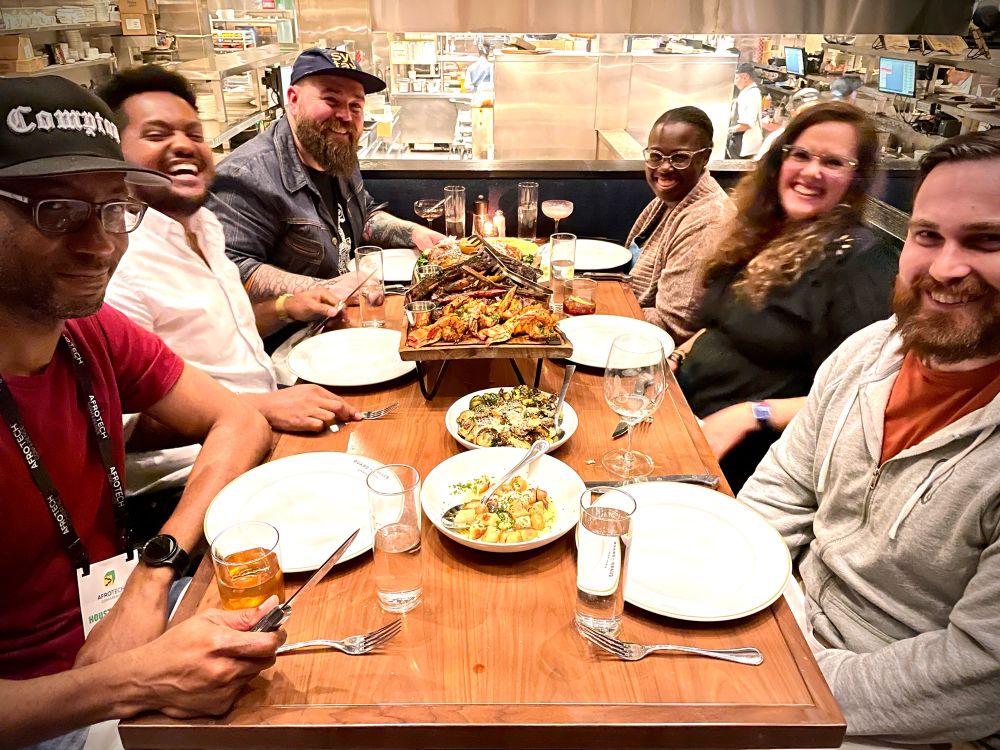 another photo of the same scene of the group at the Guard + Grace with a feast on the table. everyone has moved slightly except Anthony, who’s still mugging for the camera