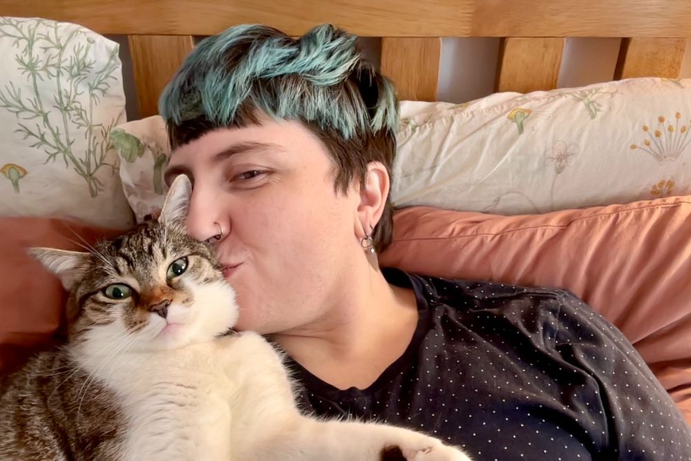 Robin lies in bed and kisses Thistle, a tabby cat who's lying on top of them and making a goofy face at the camera. 
