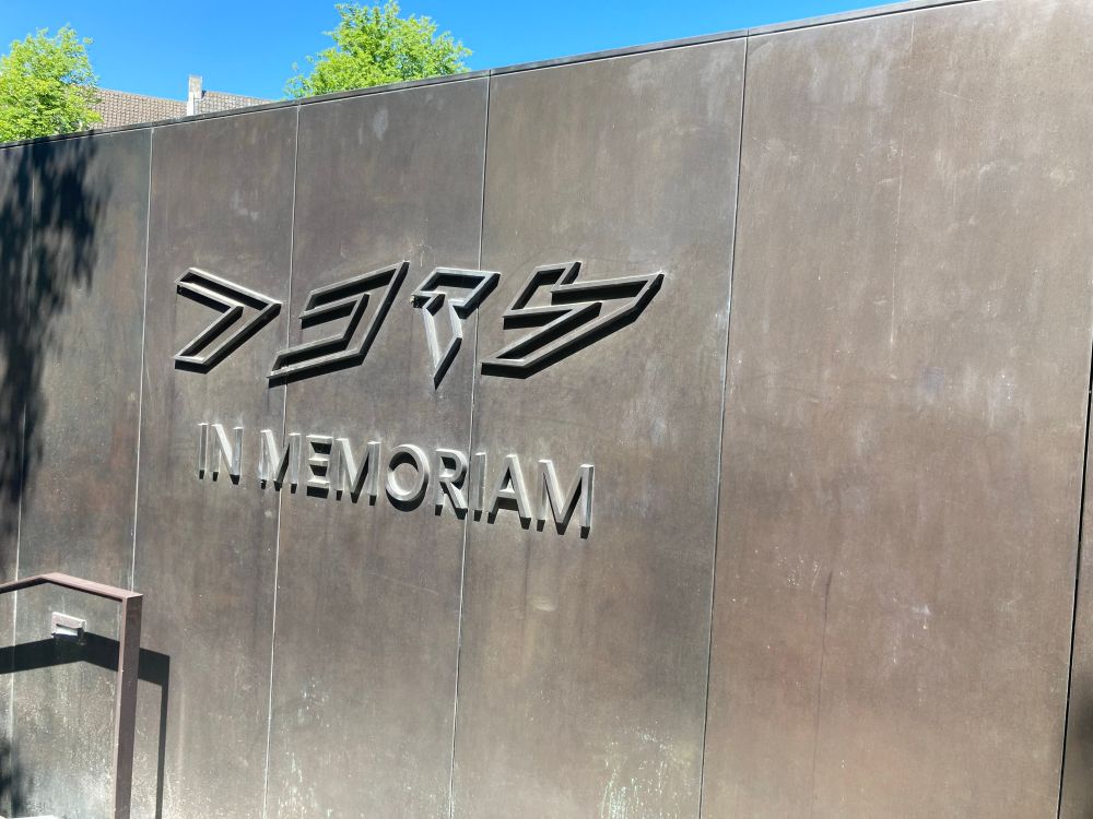 Een grijze wand waarop de tekst In Memoriam staat. Daarboven de Hebreeuwse tekst die ‘In herinnering aan’ betekent