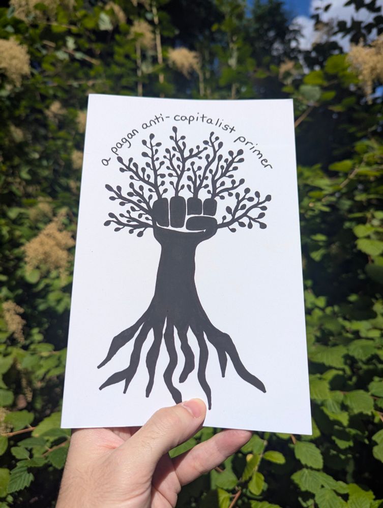 Photo of the cover of the book "A Pagan Anti-Capitalist Primer" with contrasting art in black paint of a fist with roots and leaves against a white background. I am holding the book from the bottom, in front of a large shrub.