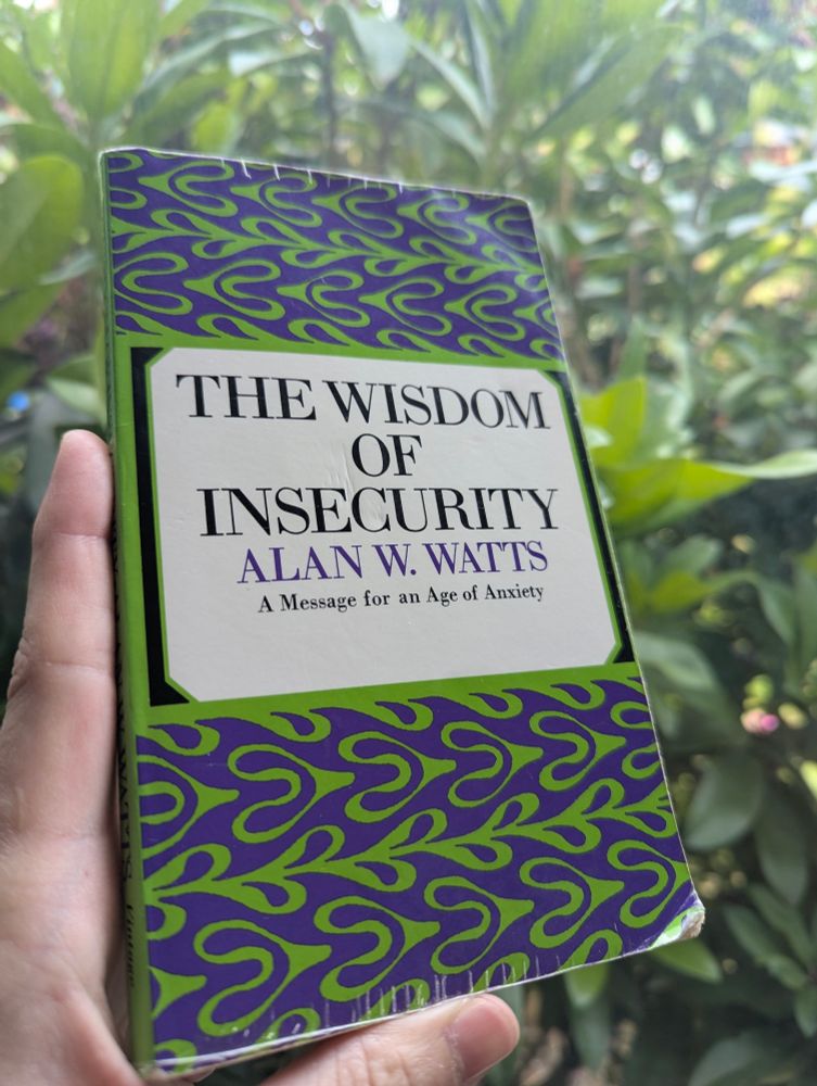 Photo of the book, "The Wisdom of Insecurity: A Message for an Age of Anxiety" by Alan W. Watts. The book cover has a slightly psychedelic pattern to it in green and purple with the title and author in the center. I am holding the book in front of a window with a rhododendron on the other side. 