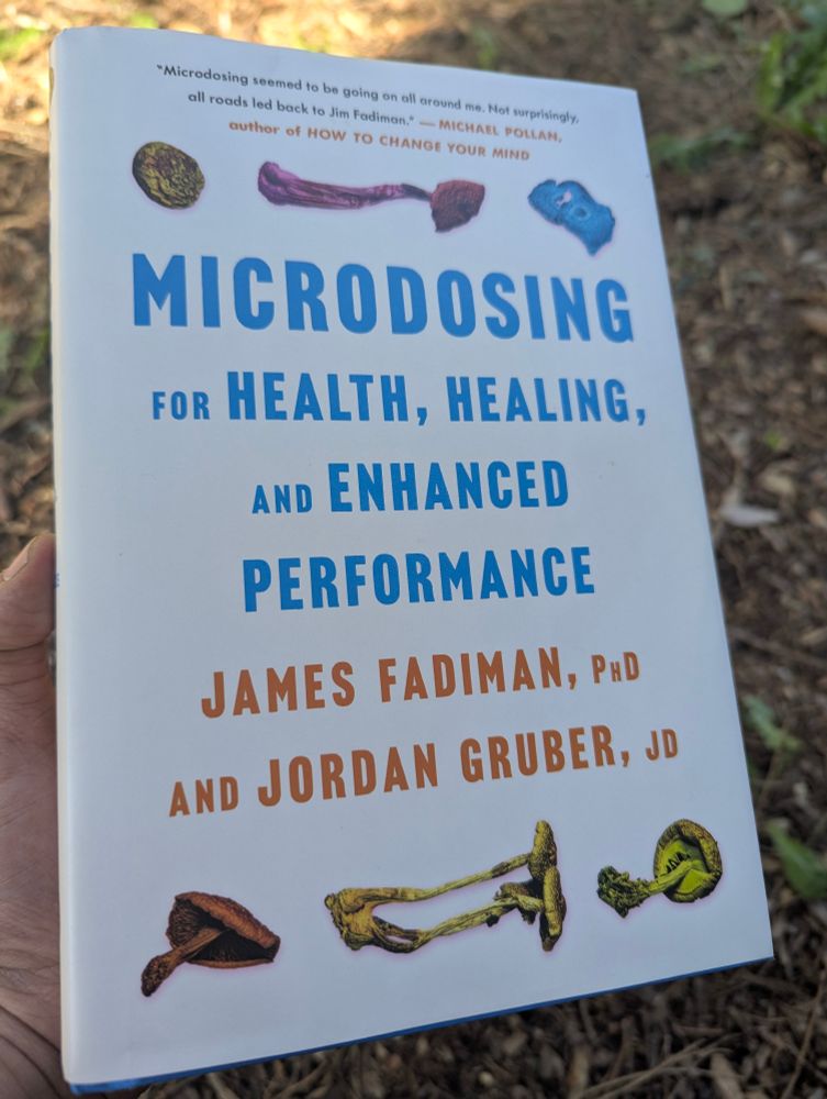 Photo of a book with a white cover and blue lettering, "Microdosing for Health, Healing, and Enhanced Performance" by James Fadiman, PhD and Jordan Gruber, JD. I am holding the book while outside, with a background of wood chips and foliage.