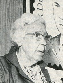 Black and white headshot of Ruth Ella Moore speaking at a podium, later in her career. She has gray hair, and is wearing glasses and pearls. 
