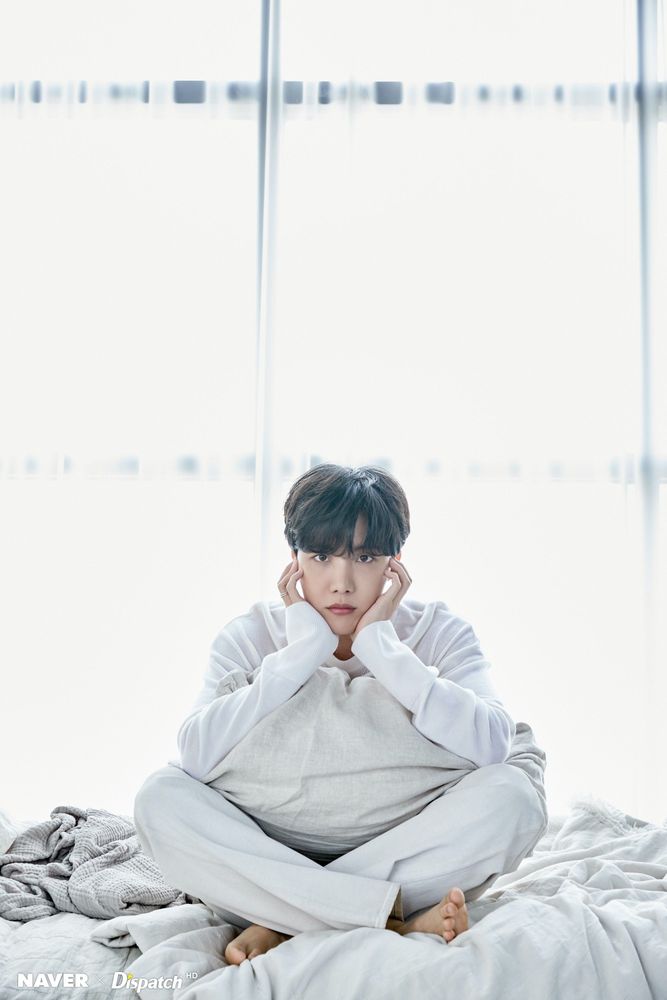 Hobi sitting cross legged on a bed. He's dressed all in white, with a pillow on his lap and his head in his hands.