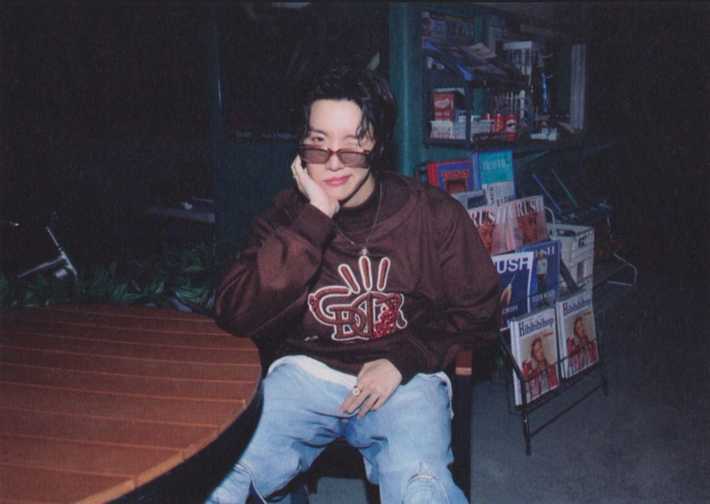 Hobi in a brown jacket and he is sitting with his elbow on a table with his head resting in his hand. 