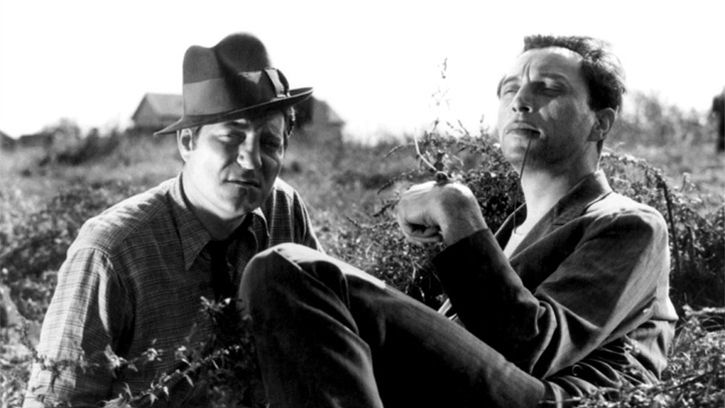 Two men relaxing in a sunny field. One has a snail perched on his hand. 