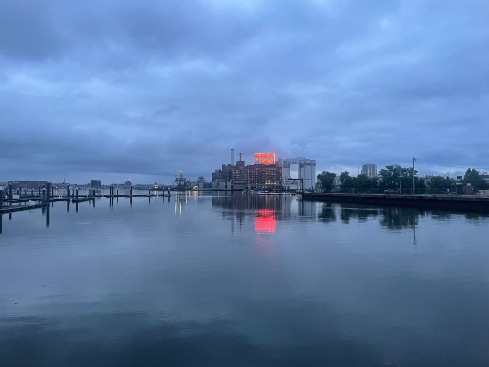 A harbor at sunrise on an overcast morning, the Domino Sugar plant in the background. 