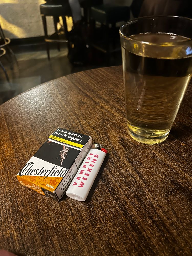 A photo of a bar table with Cigarettes, a Vampire Weekend Lighter, and a Beer