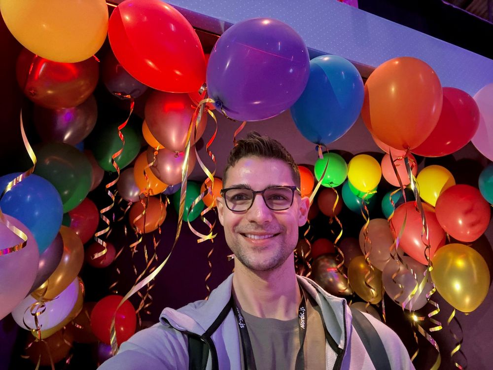 A man with glasses smiles in front of a balloons in various colors and sizes adorned with shiny ribbons.