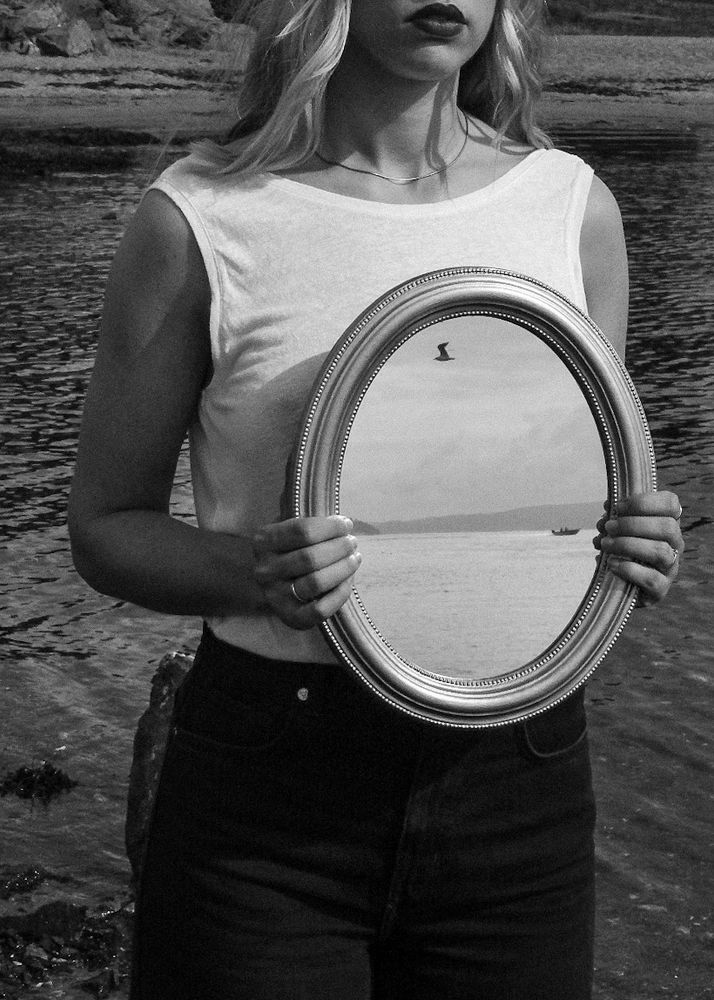 A woman standing on the beach, holding onto a mirror that reflects the ocean with a boat driving past and a bird flying in the sky. 