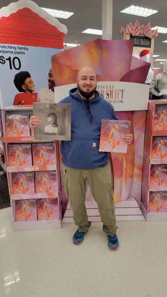 A photo of my boyfriend standing in front of Taylor Swift displays at Target holding both The Anthology vinyl and The Eras Tour book