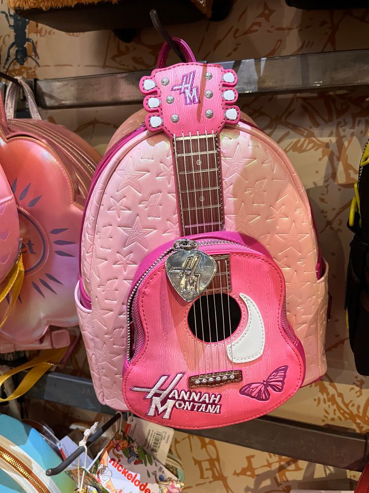 A pink Hannah Montana-themed mini backpack designed to look like a guitar. The top of the bag resembles a guitar headstock, embroidered with “HM” and six tuning pegs. The front features a guitar body shape with white detailing, faux strings, and a silver heart-shaped pick attached to the zipper. The words “Hannah Montana” are stitched in bold white and pink lettering near the bottom, along with a small embroidered butterfly. The main body of the backpack is a soft, gradient pink embossed with stars.
