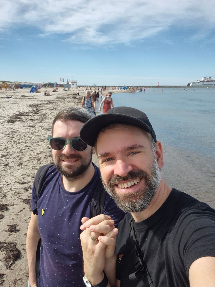 Zwei Personen halten Hänchen vor der kulisse aus Ostseestrand und einer Fähre im Hintergrund. Man sieht an einer Hand einen Ring. 