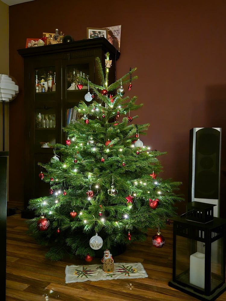 Weihnachtsbaum mit roten und weißen Kugeln geschmückt. Ein Strohengel hängt an der Baumspitze. Rechts neben dem Baum steht eine schwarze Laterne. Vor dem Baum liegt eine Weihnachtsdecke, auf der ein kleines Räuchermännchen steht.