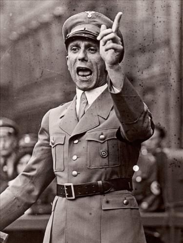 Black and white photo of Joseph Goebbels in uniform, mouth open. His arm is held up with a finger pointed, gesturing as he speaks.