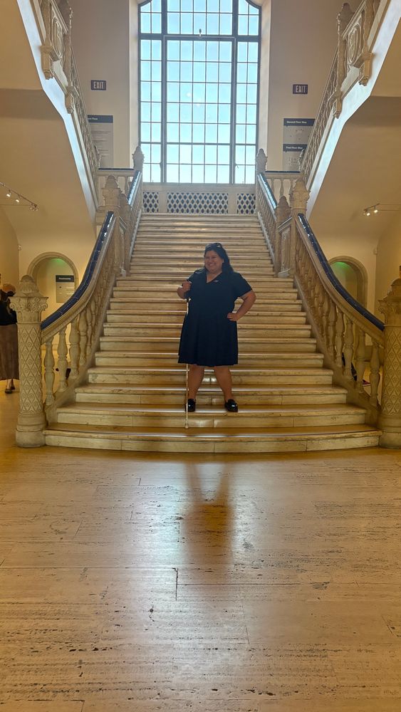 Joey, a woman wearing a blue dress and holding a white cane, smiles and stands on a staircase