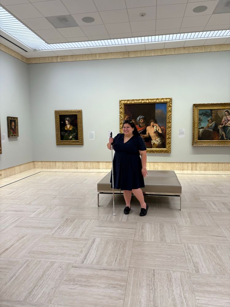 Joey, a smiling woman wearing a blue dress and holding a white cane, stands in a gallery filled with historic paintings