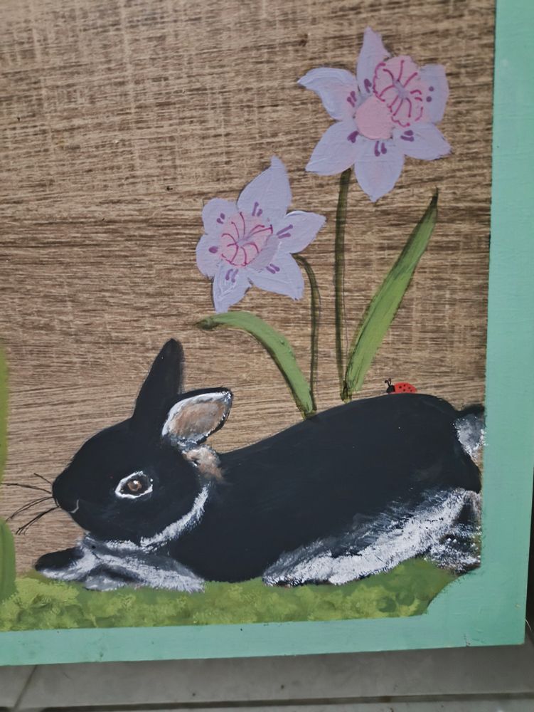Pintura sobre MDF, cor de madeira, de coelho preto e branco deitado na grama em frente a flores lilases 