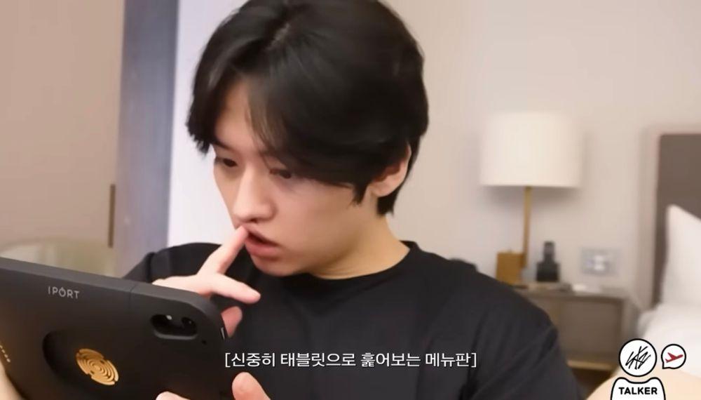 Lee Know of Stray Kids holding a tablet in a black t-shirt. He’s ordering food, pressing one finger into the top of his lip until it touches the tip of his nose