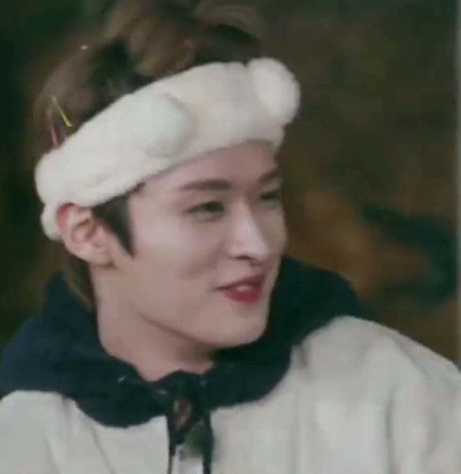lee know of stray kids in a white hoodie with a black hood. He’s wearing a fluffy white headband and smiling, only one tooth visible 