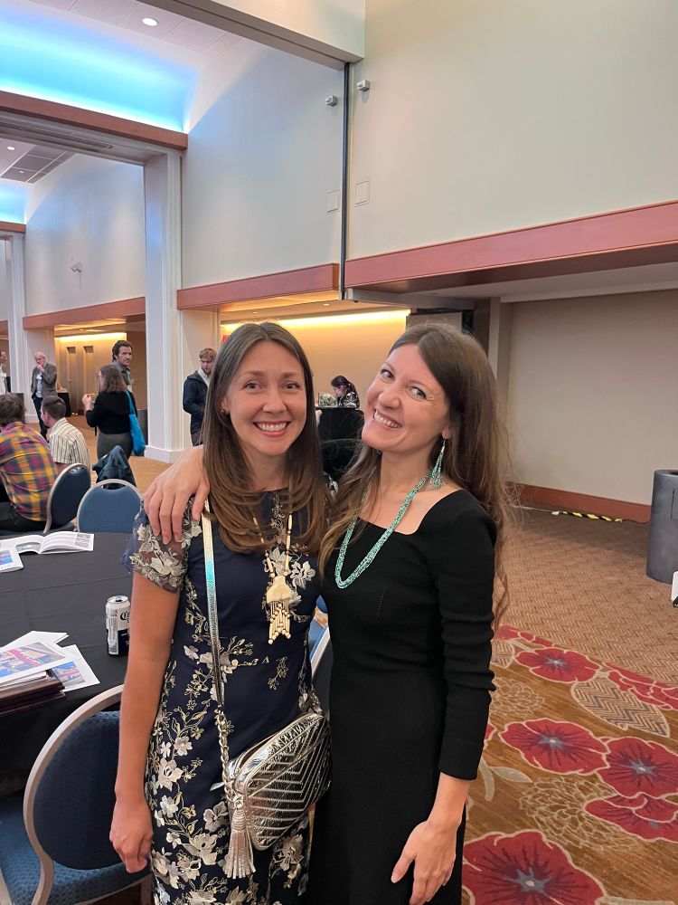 Two Native women scholars and historians, both smiling. Sarah Whitt wearing lovely turquoise jewelry a black dress, Holly Miowak Guise wearing blue embroidered dress and ivory. Conferences background with chairs and people from WHA conference 
