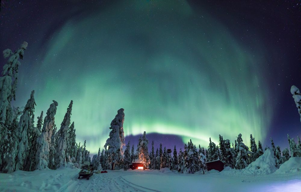 Image of the Northern Lights near Umeå, Sweden. Glowing green night sky with a small cottage and snow covered forest trees in the foreground. Photo credit: Fredrik Larsson / Visit Umeå Content Bank 