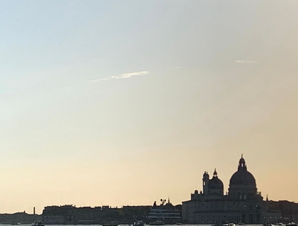 A Venice skyscape in silhouette the dome of Santa Maria della Salute prominent on the right. Hazy light blue sky.