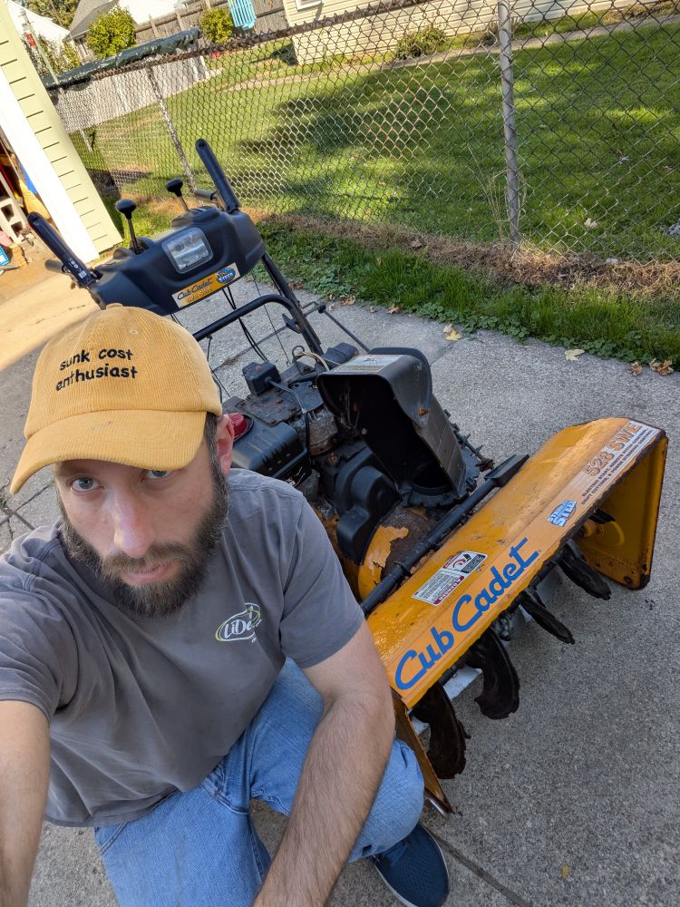 A guy with a rusty snowblower. He's wearing a hat that says "Sunk Cost Enthusiast" 