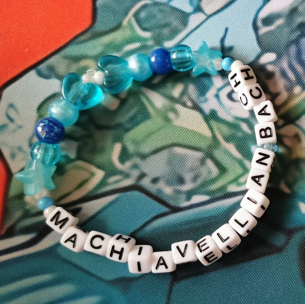 A photo of a bracelet. It has a blue and white theme, with letters that spell out "MACHIAVELLIAN BACH", a Portal 2 song reference.