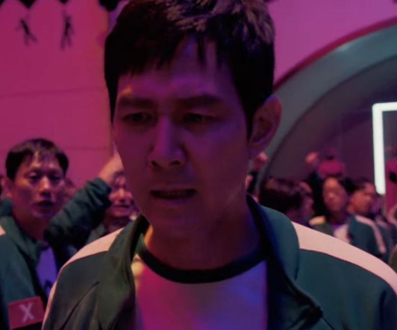 Seong Gi-Hun from Squid Game season 2, right before he yells "I've played these games before". He is a middle aged Korean man with short black hair wearing a green and white jumpsuit bathed in pink light, surrounded by a crowd of people wearing the same jumpsuit.