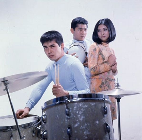Tetsuya in the foreground sitting at a drum kit. Meiko is behind him and looking at him. Tatsuya is behind her and looking at her.