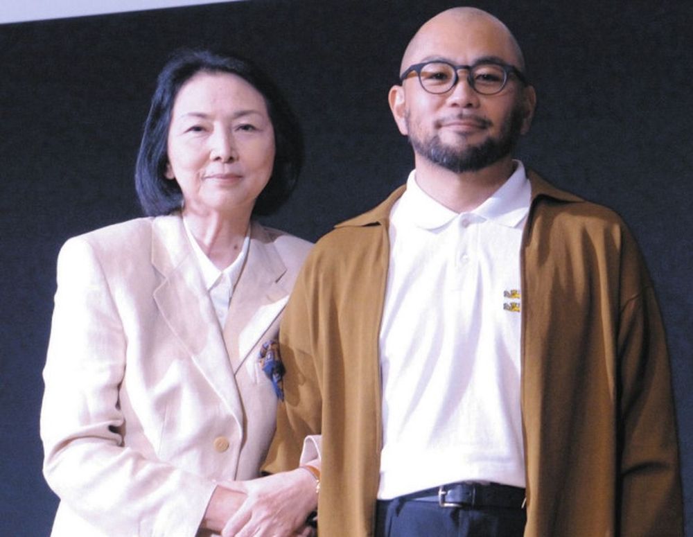Meiko Kaji and Masaaki Kudo posing for a photo on stage. It's a mid-shot, Meiko has her arm around his and they're both smiling slightly.