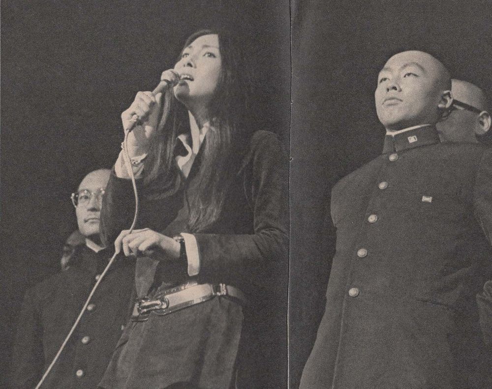 Black and white image of Meiko singing into a microphone while students stand on stage with her.