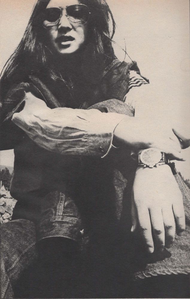 Black and white closeup of Meiko Kaji sitting down with her arms crossed in front of her knees. The camera is around foot level. She's got sunglasses on.