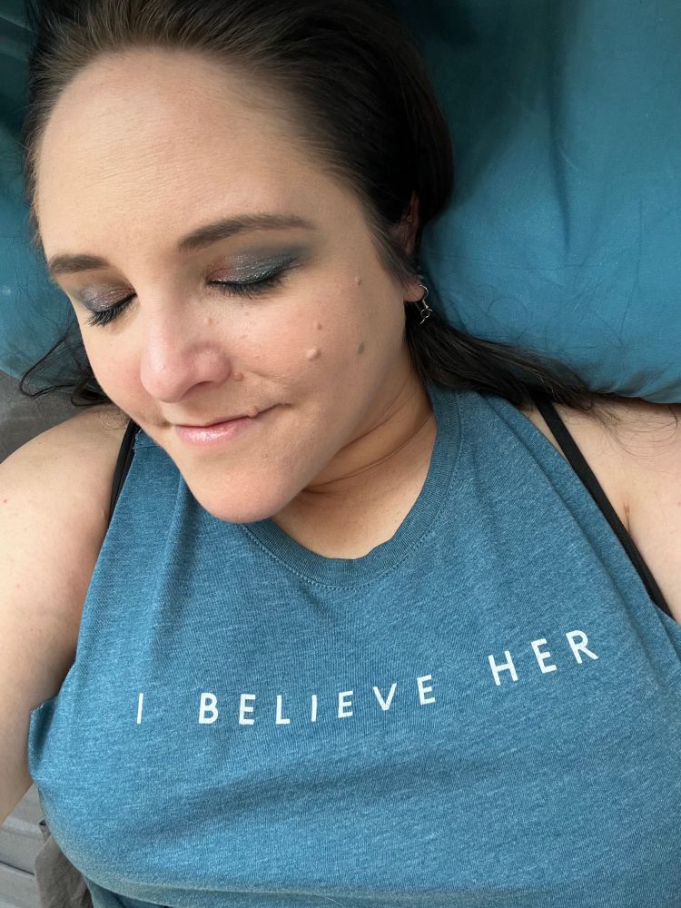 Selfie of Katie with her eyes closed to show off pink to blue eyeshadow. Wearing a teal tank top with “I believe her” printed on it. 