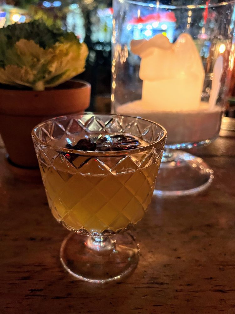 Crystal cut short-stemmed glass with a bourbon-based cocktail in it shown in front of a large half-melted white candle in a glass display on the dinner table. 