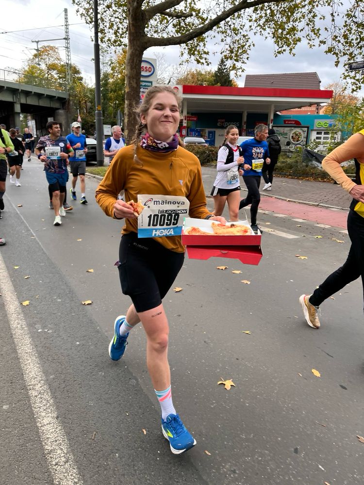 Eine junge Frau in Laufkleidung und mit Startnummer, die laufend einen Pizzakarton trägt und ein Stück der Pizza isst und lächelt. Im Hintergrund viele andere Läufer*innen. 