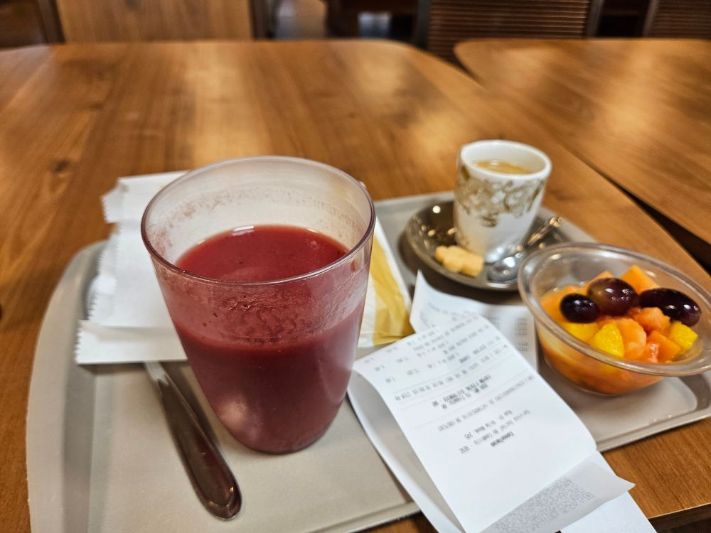 Uma bandeja contendo um salgado num saquinho de papel, um suco de morango, uma salada de frutas, café e uma nota fiscal, sobre uma mesa de madeira