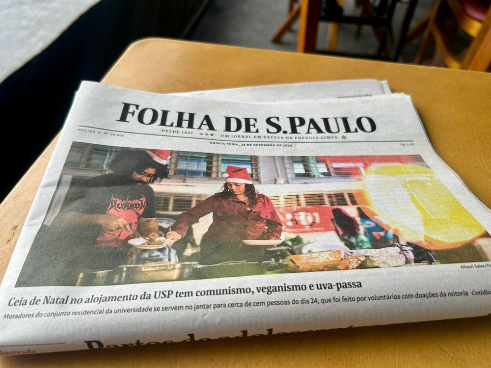 Capa da Folha de 26 de dezembro de 2024, com uma foto ocupando a metade superior da página mostrando moradores do conjunto residencial da USP se servindo de comida e usando chapéus de papai noel durante jantar no dia 24 e o título da reportagem: "Ceia de Natal no alojamento da USP tem comunismo, veganismo e uva-passa"