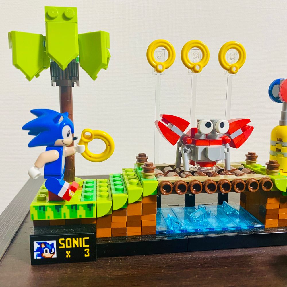 레고 ‘소닉 더 헤지혹 - 그린 힐 존’ 세트의 왼쪽 끝부분을 찍은 사진. 메가드라이브 게임 ‘소닉 더 헤지혹’의 첫 스테이지 ‘그린 힐 존’을 재현한 블럭 조립 세트입니다. 짙고 연한 갈색 체크무늬 지형에 푸른 풀밭, 야자수와 개울에 걸친 통나무 다리 등의 배경 요소를 레고 블럭으로 충실히 재현했으며, 공중에 떠 있는 링도 투명 막대에 끼우는 형식으로 재현. 왼쪽 아래 끝 부분에는 게임에도 있었던 플레이어 수 표시 ‘SONIC X 3’이 보입니다. 오리지널 미니피규어로 재현된 소닉이 한 손에 링을 들고 지금이라도 달려갈 듯한 포즈를 취하고 있고, 통나무 다리 위에는 게 형태의 적 로봇이 길을 막고 있습니다.