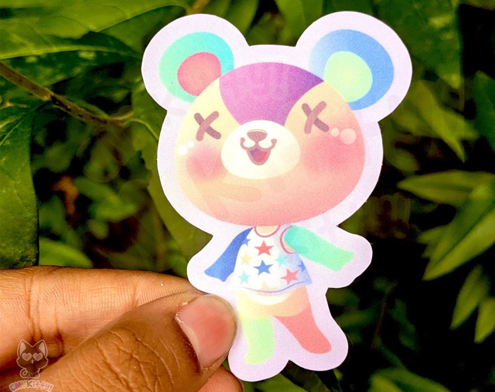 I am holding a sticker I designed of Stitches the cub from Animal Crossing. He is looking very happy! There are bushes behind him. 