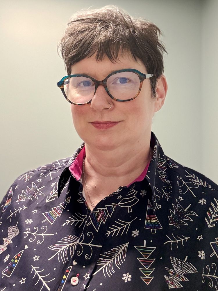 Woman with short brown hair and glasses wearing a black shirt with white trees on it, with some trees having rainbow colors in them. 
