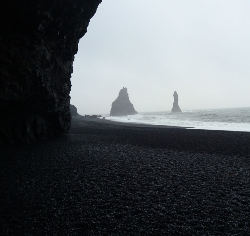View of the sea across a black sand beach, taken from just inside a basalt cave. The sky is pale grey, the sea a slightly darker grey with white crested waves. 2 large dark rock formations can be seen in the sea through the mist.