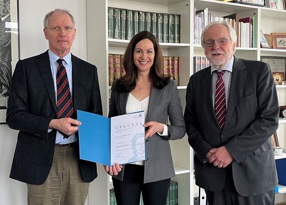Three people stand in an office setting with bookshelves in the background. On the left, Prof. Dr. Stefan Kadelbach, Dean of the Faculty of Law, wearing a dark suit and striped tie. Together with Mateja S. Platise in the center, wearing a gray blazer over a white top, they hold a certificate in a blue folder. On the right, Prof. Dr. Reiner Hofmann, wearing a dark suit, glasses, and striped tie, stands with hands clasped. The occasion is Platise's appointment as Honorary Professor at Goethe University Frankfurt.