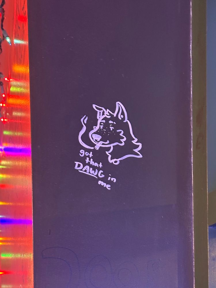 white paint doodle on a black wall. it’s a headshot of an anthropomorphic, shepherd-like dog, with one pointed ear and one ear half flopped over. he has ear gauges and a septum ring, and is smoking a joint. “got that DAWG in me” is written in the bottom left corner of the doodle.
