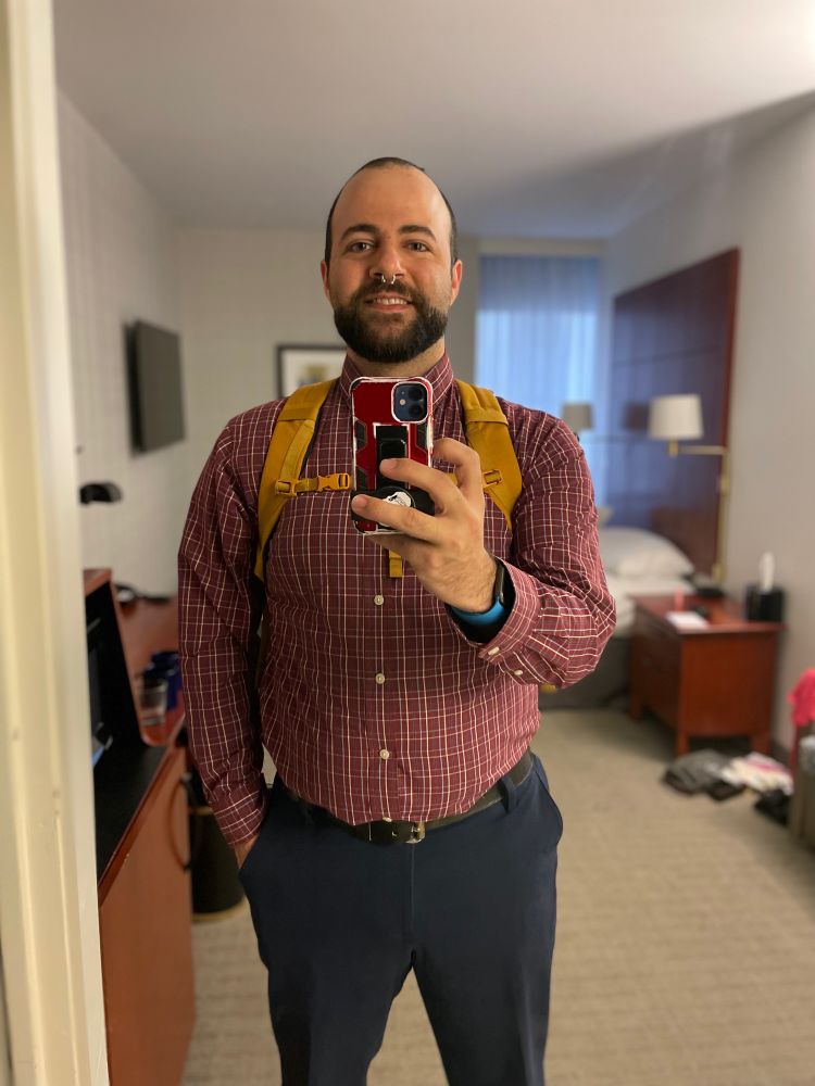 Bearded person with a backpack wearing a shirt and pants in a hotel room. 