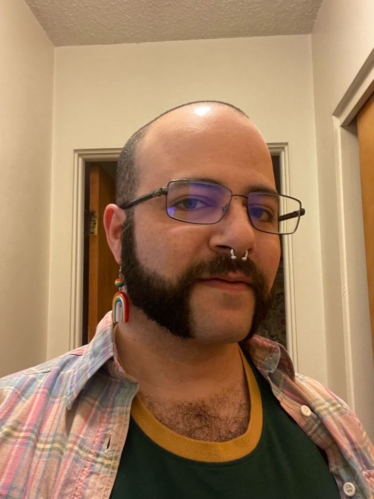 A selfie of Tony wearing a light pink and light blue plaid shirt with a Smokey the Bear t-shirt under. Tony is wearing glasses, rainbow earrings made from clay, and a 10-gauge septum ring.
