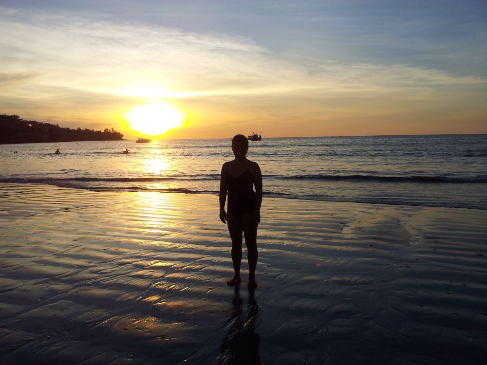 Eine Frau steht mit den Füßen im Wasser am Strand in der Abenddämmerung und blickt auf die untergehende Sonne am Meer in Koh Samui Thailand.