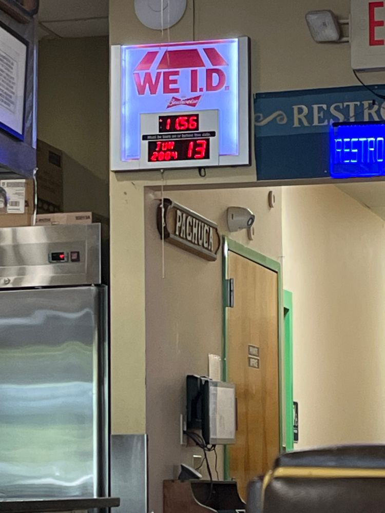 A sign in a restaurant that says “WE I.D.” above the Budweiser logo. A digital clock below that says 11:56. Then “Must be born on or before this date, June 13, 2004.” The date is displayed digitally. 