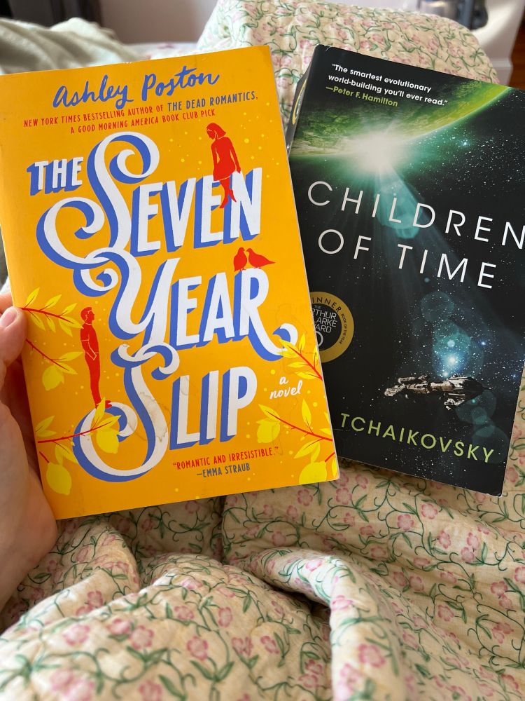 A hand holding two books. On the left, the seven year slip with a yellow cover that yellow leaves and a couple in red looking at each other. On the right, children of time, which has a cover with a green planet and a spaceship orbiting the planet.
